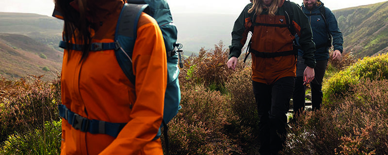 Vrouw en twee mannen wandelen door de natte, heuvelachtige bergen in herfst/winter kleding, met mutsen en wandelrugzakken. De vrouw draagt een oranje jas, man 1 een bruine en zwarte jas, en man 2 een donkerblauwe jas. Het pad is nat en de omgeving is grijs en grauw